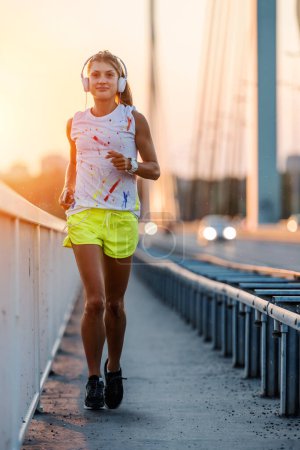 Foto de Mujer joven atleta con auriculares corriendo por el puente en la puesta del sol - Imagen libre de derechos