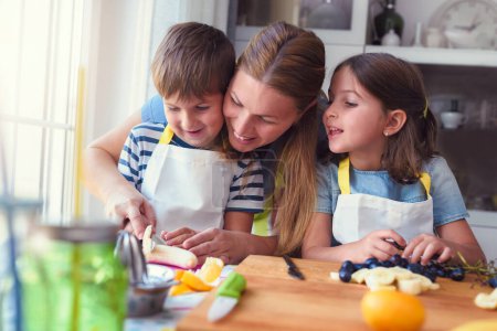 Foto de Niños pequeños y felices con su madre en la cocina - preparando un bocadillo de frutas saludables - Imagen libre de derechos