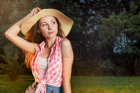 Photo for Pretty Fashion Country Girl with Summer Hat Sitting on Fence Outdoors - Royalty Free Image