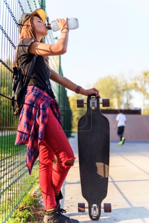 Téléchargez les photos : Jolie fille urbaine tenant longboard dans un skate park avec bouteille d'eau - en image libre de droit