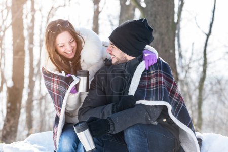 Foto de Pareja joven sonriente con copas sentadas en el bosque de montaña cubierto de nieve. Beber bebidas calientes, divertirse. - Imagen libre de derechos