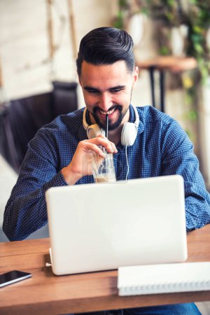 Téléchargez les photos : Joyeux jeune homme avec barbe travaillant sur ordinateur portable dans un café - en image libre de droit