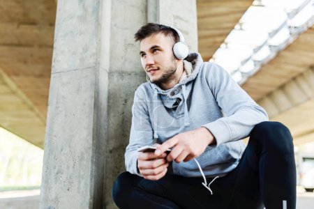 Photo for Urban handsome man runner with earphones taking break under bridge listening music - Royalty Free Image