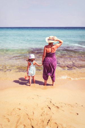Téléchargez les photos : Vue arrière d'une mère et d'une fille debout sur une plage d'émeraude parfaite - en image libre de droit