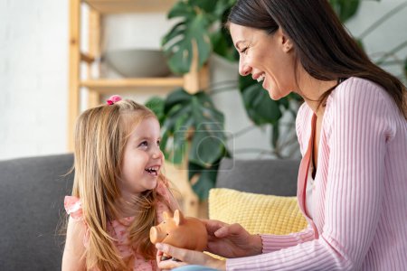 Photo for Young mother holding piggy bank while sitting at home with cute daughter - Royalty Free Image