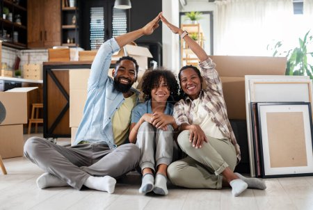 Foto de Familia afroamericana feliz sentado en el suelo en un nuevo hogar, mirando a la cámara, cajas móviles con nueva casa y cajas de cartón, se mueve en un nuevo apartamento - Imagen libre de derechos