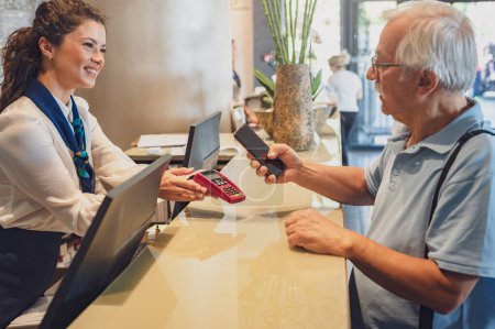 Téléchargez les photos : Un couple de personnes âgées à la réception de l'hôtel effectue des paiements sans contact avec un smartphone. Les aînés voyageant et utilisant les nouvelles technologies - en image libre de droit