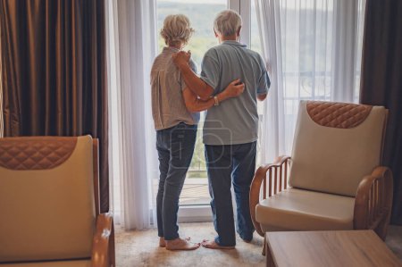 Photo for An elderly married couple just arrived at the hotel, looking through the window of their luxury apartment - Royalty Free Image