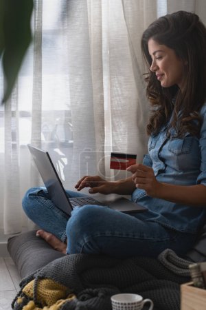 Téléchargez les photos : Belle jeune femme souriante assise confortablement près de la fenêtre à la maison, en utilisant l'ordinateur pour acheter des cadeaux de vacances en ligne avec une carte de crédit - en image libre de droit