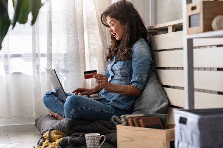 Téléchargez les photos : Belle jeune femme souriante assise confortablement près de la fenêtre à la maison, en utilisant l'ordinateur pour acheter des cadeaux de vacances en ligne avec une carte de crédit - en image libre de droit