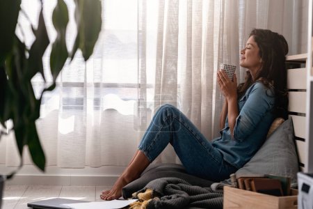 Téléchargez les photos : Belle jeune femme souriante assise à la maison dans un coin de fenêtre confortable, buvant du café - en image libre de droit