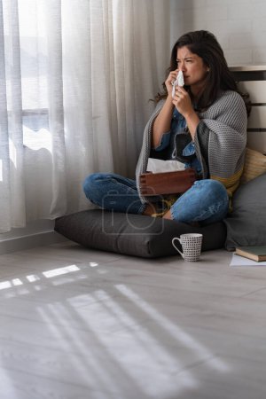 Téléchargez les photos : Jeune femme à la maison essuyant son nez, éternuant, ayant un rhume, la grippe, les allergies. Essayer de se réchauffer. - en image libre de droit