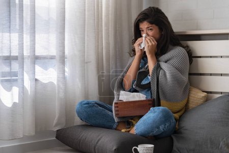 Foto de Mujer joven en casa limpiándose la nariz, estornudando, resfriada, gripe, alergias. Tratando de calentar. - Imagen libre de derechos
