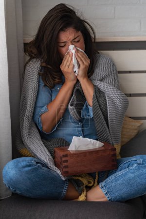 Téléchargez les photos : Jeune femme à la maison essuyant son nez, éternuant, ayant un rhume, la grippe, les allergies. Essayer de se réchauffer. - en image libre de droit