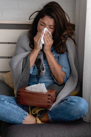 Photo for Young woman at home wiping her nose, sneezing, having a cold, flu, allergies. Trying to warm up. - Royalty Free Image