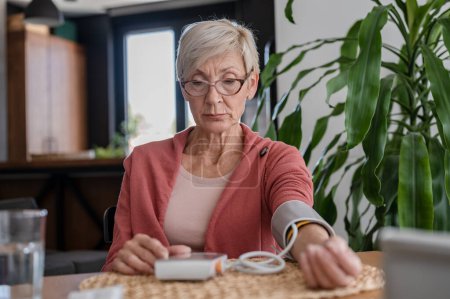 Téléchargez les photos : Femme âgée assise seule à la maison, mesurant sa tension artérielle avec un appareil domestique - en image libre de droit