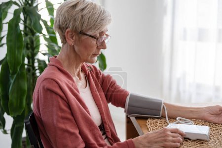 Téléchargez les photos : Femme âgée assise seule à la maison, mesurant sa tension artérielle avec un appareil domestique - en image libre de droit