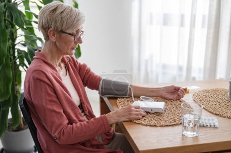 Foto de Mujer mayor sentada sola en casa, midiendo su presión arterial con un dispositivo casero - Imagen libre de derechos