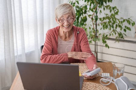 An elderly woman sitting in front of a laptop computer at home, having an online video call with a doctor. Online medical consultation
