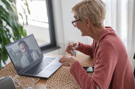 Téléchargez les photos : Une femme âgée assise devant un ordinateur portable à la maison, ayant un appel vidéo en ligne avec un médecin. Consultations médicales en ligne. - en image libre de droit