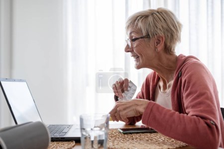 Una anciana sentada frente a una computadora portátil en casa, recibiendo una videollamada en línea con un médico. Consulta médica en línea