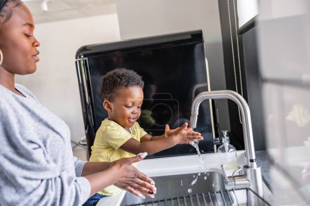 Photo for Cute little smiling African-American boy washing hands in a kitchen sink at home - Royalty Free Image