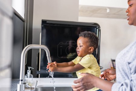 Téléchargez les photos : Mignon petit garçon afro-américain souriant se lave les mains dans un évier de cuisine à la maison - en image libre de droit