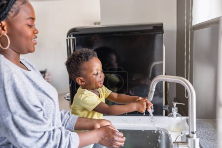 Photo for Cute little smiling African-American boy washing hands in a kitchen sink at home - Royalty Free Image