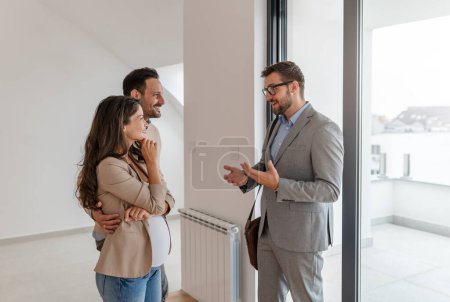 Téléchargez les photos : Homme et sa femme enceinte, parlant avec un agent immobilier visitant un appartement à vendre ou à louer. Les futurs parents achètent un appartement. Concept immobilier - en image libre de droit