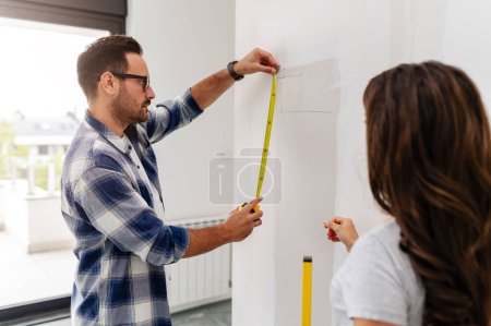 Photo for A young, beautiful, and cheerful couple, comprising a man and his pregnant wife, are measuring the walls, as part of their new home renovation project - Royalty Free Image