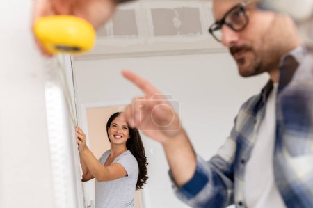 Photo for A young, beautiful, and cheerful couple, comprising a man and his pregnant wife, are measuring the walls, as part of their new home renovation project - Royalty Free Image