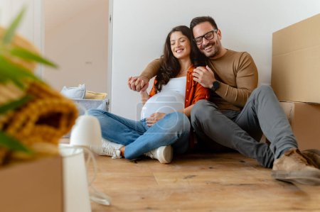 Photo for Young pregnant woman and her husband with cardboard boxes sitting on the floor. - Royalty Free Image