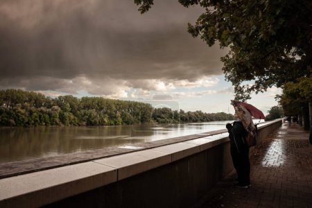 Photo for SZEGED, HUNGARY - SEPTEMBER 18, 2022: Selective blur on two women with umbrellas standing in front of the tisza river at dusk, during a rainy evening, in the riverbank of Szeged, a hungarian city. - Royalty Free Image