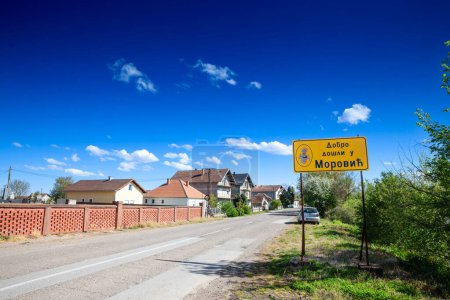 Foto de MOROVIC, SERBIA - 25 DE ABRIL DE 2022: Señal de tráfico que indica la entrada a Morovic con una carretera rural local. Morovic es un pueblo serbio en el oeste de Serbia, cerca de la ciudad de Sid, en la región de Srem, en verano, en Voivodina.. - Imagen libre de derechos