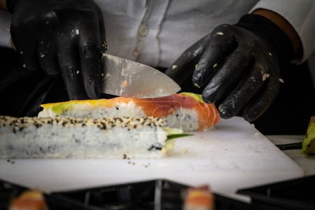 Foto de Desenfoque selectivo en las manos de un chef que prepara y corta sushi y sashimi hechos de salmón con un cuchillo, preparándose para ser entregados en un sushi para llevar. - Imagen libre de derechos