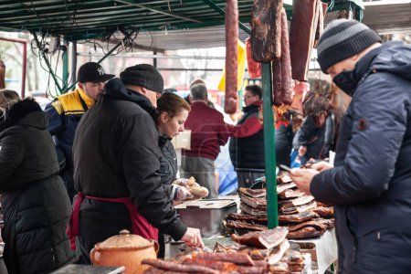 Photo for KACAREVO, SERBIA - MARCH 6, 2022: Selective blur on a Couple of butchers selliing smoked & cured meat, sausage, kosasica style, to clients on a stand at the Slaninijada Kacarevo market in the serbian countryside. - Royalty Free Image
