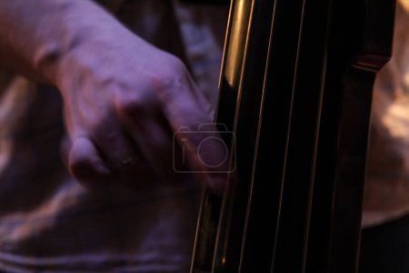 Téléchargez les photos : Flou sélectif sur les mains d'un musicien touchant les cordes d'un violoncelle contrebasse, jouant du pizzicato, dans un environnement sombre, avec un mouvement de vitesse dû aux mains se déplaçant et jouant de l'instrument de musique. - en image libre de droit