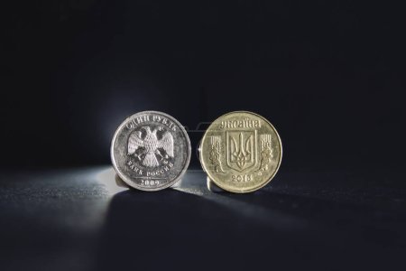 Selective blur on a one ruble coin with the mention one ruble and bank of russia written in russian, next to a ukrainian hryvnia coin with Ukraine written in ukrainian isolated on a black background.