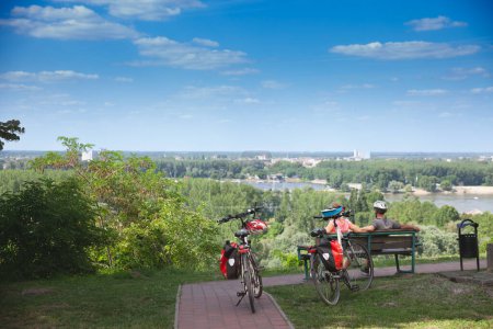 Téléchargez les photos : ILOK, CROATIE - 18 JUIN 2022 : Flou sélectif sur un couple avec vélo faisant une pause pendant une balade à vélo à Ilok, regardant le fleuve Danube, avec des bagages sur le vélo, lors d'un voyage touristique d'été. - en image libre de droit