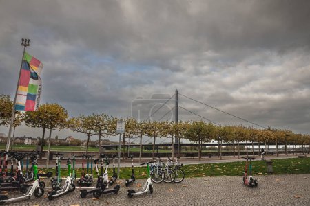 Téléchargez les photos : DUSSELDORF, ALLEMAGNE - 7 NOVEMBRE 2022 : Panorama d'une station de partage avec scooters électriques et vélos à louer devant le pont oberkasseler brucke à l'automne à Düsseldorf. - en image libre de droit