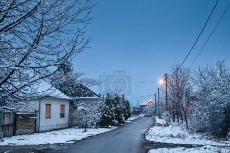 Téléchargez les photos : Flou sélectif sur une route et une rue vides dans le village de Bavaniste, en Voïvodine, Banat, Serbie, à la campagne, couvert de neige, pendant une nuit froide et glacée crépuscule d'hiver. - en image libre de droit