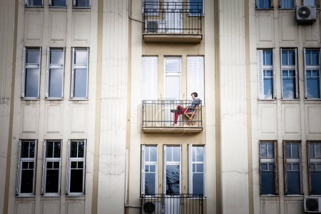 Téléchargez les photos : BELGRADE, SERBIE - 30 AVRIL 2023 : Homme, travailleur masculin en ligne, nomade numérique, pigiste, travaillant de chez lui assis sur son balcon à Belgrade, pendant un après-midi ensoleillé, avec son ordinateur portable. - en image libre de droit