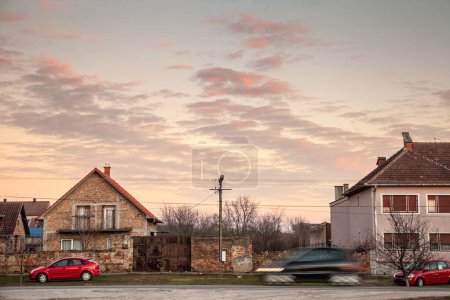 Téléchargez les photos : Focus sélectif sur les maisons résidentielles dans le village serbe de Banatsko Novo Selo avec la voiture conduisant rapidement avec un flou de vitesse passant devant elle sur une route locale. c'est un paysage rural de Serbie & Europe. - en image libre de droit