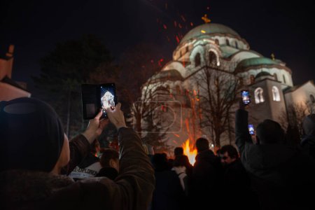 Foto de BELGRADE, SERBIA - 6 DE ENERO DE 2023: Desenfoque selectivo en un hombre que toma una foto con su teléfono inteligente del pueblo serbio quemando ramas de bádnjak durante la Navidad ortodoxa frente a la catedral de San Sava. - Imagen libre de derechos