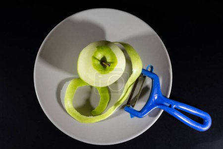 Téléchargez les photos : Flou sélectif sur une pomme verte à moitié pelée avec une écorce de pomme sur une assiette grise isolée sur un fond noir avec un éplucheur de légumes et de fruits, un éplucheur Y. - en image libre de droit