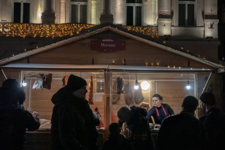 Téléchargez les photos : NOVI SAD, SERBIE - 4 DÉCEMBRE 2022 : Flou sélectif sur les Boutiques du marché de Noël de Novi Sad avec des gens qui achètent des choses dans des hangars et des étals d'artisans la nuit, sur la place trg slobode. - en image libre de droit