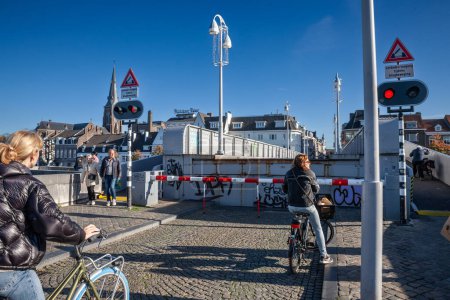 Foto de MAASTRICHT, PAÍSES BAJOS - 10 DE NOVIEMBRE DE 2022: Ciclistas esperando frente al puente Sint servaasbrug, o puente de san servatius, el puente más antiguo de los Países Bajos, un símbolo de puente elevador de maastricht. - Imagen libre de derechos