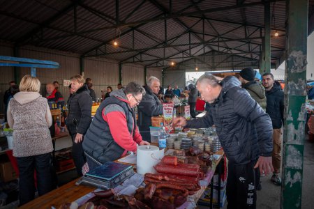 Photo for RUMENKA, SERBIA - JANUARY 28, 2024: Stand of a butcher in the Pihtijada Rumeka market selling sausages and cured meat, with a client buying products of Serbian agriculture. - Royalty Free Image