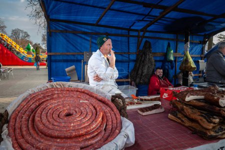 Photo for KACAREVO, SERBIA - FEBRUARY 18, 2023: Stand of a butcher in the Slaninijada Kacarevo market selling sausages, with a butcher smoking cigarettes in front. - Royalty Free Image