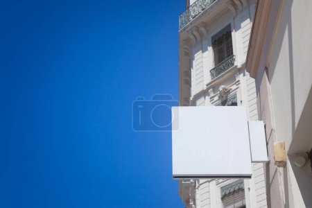 Selective blur on a Empty white square shop signboard hanging on a building facade, visible against a clear sky in a French city street ideal for retail business advertising & urban commercial display.